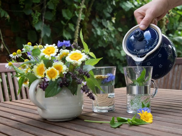 hand giesst wasser aus teekanne in glas neben blumenstrauss